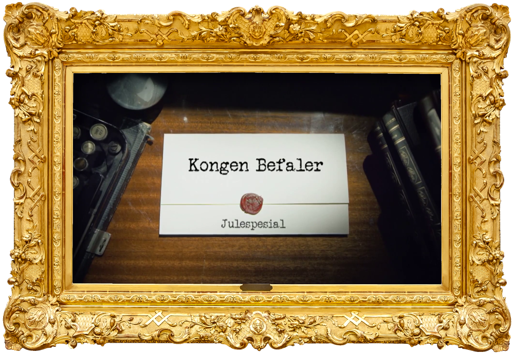 Image of the title card at the start of this episode, showing a task brief with the show title, 'Kongen Befaler', and the episode title, 'Julespesial' ['Christmas special'], on a wooden desk. At the edges of the image, part of a typewriter keyboard and a stack of leather-bound books can be seen.