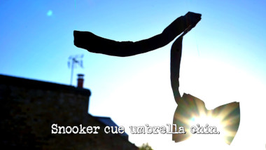 Photo of a bowtie suspended in mid-air, silhouetted against the sun, in front of the Taskmaster house (referencing the 'Take the most dramatic photo of yourself in mid-air' and 'Tie yourself to the bed' tasks), with the episode title, 'Snooker cue umbrella chin', superimposed on it.