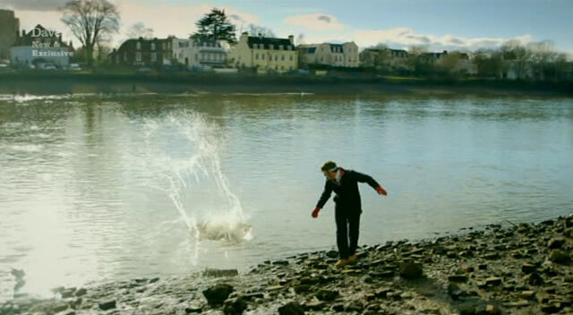 Image of Tim Key stumbling away from the river as the ice block lands in the water with a  splash.