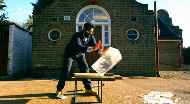 Image of Romesh Ranganathan shoving a large block of ice off of a small table in front of the Taskmaster house.