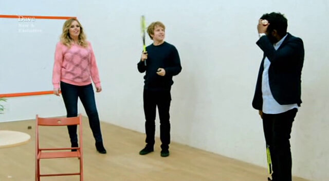 Image of Roisin Conaty, Josh Widdicombe, and Romesh Ranganathan standing in a squash court, trying to figure out how they are scoring points, while Romesh scores a point by touching his own head.