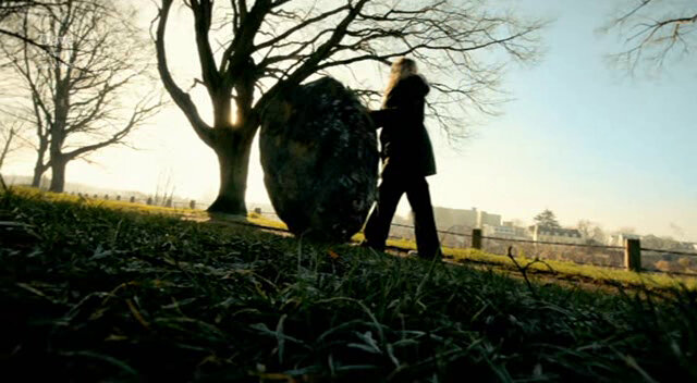 Silhouetted image of Roisin Conaty rolling a prop boulder along the river bank.