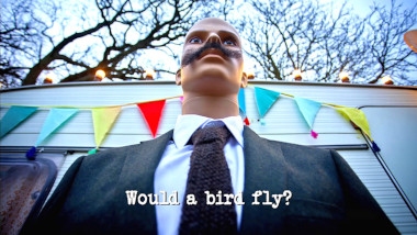 Photo taken from a low angle, looking up at a mannequin with a moustache, wearing a suit, and standing in front of the caravan (referencing the 'Deliver a medium mocha to Mike’s mate Mark' task), with the episode title, 'Would a bird fly?', superimposed on it.