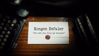 Image of the title card at the start of this episode, showing a task brief with the show title, 'Kongen Befaler', and the episode title, 'Gni det inn, folk på trikken' ['Rub it in, people on the tram'], on a wooden desk. At the edges of the image, part of a typewriter keyboard and a stack of leather-bound books can be seen.