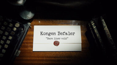Image of the title card at the start of this episode, showing a task brief with the show title, 'Kongen Befaler', and the episode title, 'Barn liker vold' ['Children like violence'], on a wooden desk. At the edges of the image, part of a typewriter keyboard and a stack of leather-bound books can be seen.