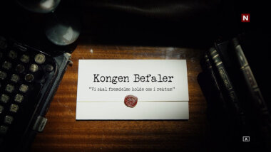 Image of the title card at the start of this episode, showing a task brief with the show title, 'Kongen Befaler', and the episode title, 'Vi skal fremdeles holde oss i rektum' ['We will still stay in the rectum'], on a wooden desk. At the edges of the image, part of a typewriter keyboard and a stack of leather-bound books can be seen.
