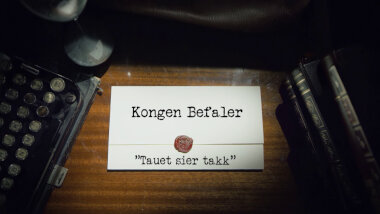 Image of the title card at the start of this episode, showing a task brief with the show title, 'Kongen Befaler', and the episode title, 'Tauet sier takk' ['The rope says thank you'], on a wooden desk. At the edges of the image, part of a typewriter keyboard and a stack of leather-bound books can be seen.