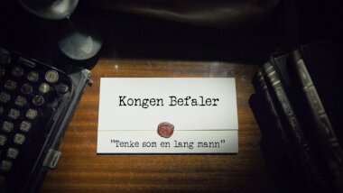 Image of the title card at the start of this episode, showing a task brief with the show title, 'Kongen Befaler', and the episode title, 'Tenke som en lang mann' ['Think like a tall man'], on a wooden desk. At the edges of the image, part of a typewriter keyboard and a stack of leather-bound books can be seen.
