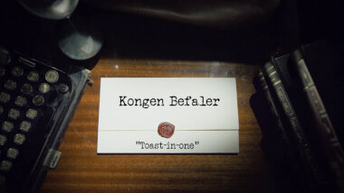 Image of the title card at the start of this episode, showing a task brief with the show title, 'Kongen Befaler', and the episode title, 'Toast-in-one', on a wooden desk. At the edges of the image, part of a typewriter keyboard and a stack of leather-bound books can be seen.