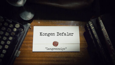 Image of the title card at the start of this episode, showing a task brief with the show title, 'Kongen Befaler', and the episode title, 'Langrennslys' ['Cross-country lights'], on a wooden desk. At the edges of the image, part of a typewriter keyboard and a stack of leather-bound books can be seen.