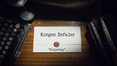 Image of the title card at the start of this episode, showing a task brief with the show title, 'Kongen Befaler', and the episode title, 'Knipestag' [a made-up word], on a wooden desk. At the edges of the image, part of a typewriter keyboard and a stack of leather-bound books can be seen.