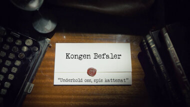 Image of the title card at the start of this episode, showing a task brief with the show title, 'Kongen Befaler', and the episode title, 'Underhold oss, spis kattemat' ['Entertain us, eat cat food'], on a wooden desk. At the edges of the image, part of a typewriter keyboard and a stack of leather-bound books can be seen.