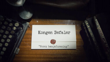 Image of the title card at the start of this episode, showing a task brief with the show title, 'Kongen Befaler', and the episode title, 'Norsk bæsjeforening' ['Norwegian pooping association'], on a wooden desk. At the edges of the image, part of a typewriter keyboard and a stack of leather-bound books can be seen.