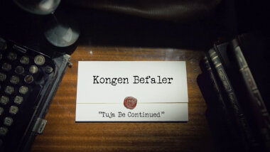 Image of the title card at the start of this episode, showing a task brief with the show title, 'Kongen Befaler', and the episode title, 'Tuja be continued', on a wooden desk. At the edges of the image, part of a typewriter keyboard and a stack of leather-bound books can be seen.