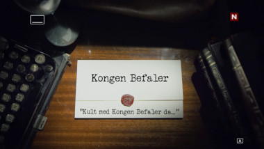 Image of the title card at the start of this episode, showing a task brief with the show title, 'Kongen Befaler', and the episode title, 'Kult med Kongen Befaler da...' ['Cool to be back with Kongen Befaler then...'], on a wooden desk. At the edges of the image, part of a typewriter keyboard and a stack of leather-bound books can be seen.