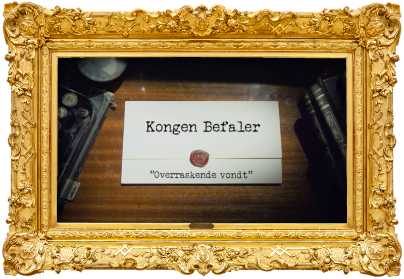 Image of the title card at the start of this episode, showing a task brief with the show title, 'Kongen Befaler', and the episode title, 'Overraskende vondt' ['Surprisingly painful'], on a wooden desk. At the edges of the image, part of a typewriter keyboard and a stack of leather-bound books can be seen.