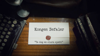 Image of the title card at the start of this episode, showing a task brief with the show title, 'Kongen Befaler', and the episode title, 'Ta deg en slurk spett' ['Take a sip of digging bar'], on a wooden desk. At the edges of the image, part of a typewriter keyboard and a stack of leather-bound books can be seen.