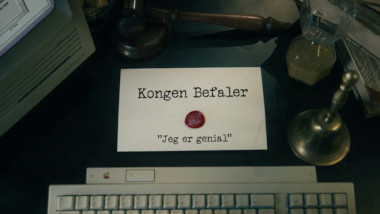 Image of the title card at the start of this episode, showing a task brief with the show title, 'Kongen Befaler', and the episode title, 'Jeg er genial' ['I'm brilliant'], on a wooden desk. Also on the desk is an old-fashioned Apple computer and keyboard, a gavel, an hourglass, and a bell.