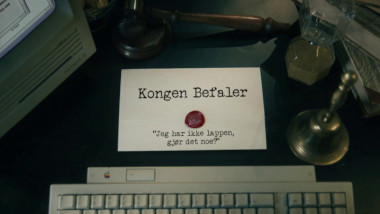 Image of the title card at the start of this episode, showing a task brief with the show title, 'Kongen Befaler', and the episode title, 'Jeg har ikke lappen, gjør det noe?' ['I don't have a license, is that okay?'], on a wooden desk. Also on the desk is an old-fashioned Apple computer and keyboard, a gavel, an hourglass, and a bell.