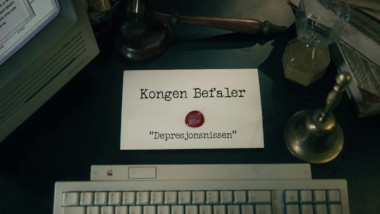 Image of the title card at the start of this episode, showing a task brief with the show title, 'Kongen Befaler', and the episode title, 'Depresjonsnissen' ['The depression gnome'], on a wooden desk. Also on the desk is an old-fashioned Apple computer and keyboard, a gavel, an hourglass, and a bell.