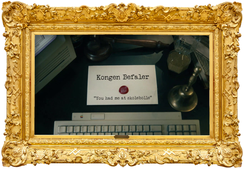 Image of the title card at the start of this episode, showing a task brief with the show title, 'Kongen Befaler', and the episode title, 'You had me at skolebolle' ['You had me at school bun'], on a wooden desk. Also on the desk is an old-fashioned Apple computer and keyboard, a gavel, an hourglass, and a bell.