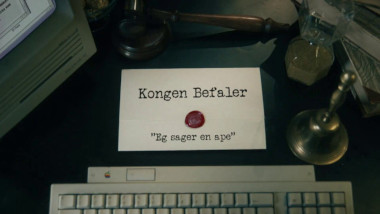 Image of the title card at the start of this episode, showing a task brief with the show title, 'Kongen Befaler', and the episode title, 'Eg sager en ape' ['I'm sawing a monkey'], on a wooden desk. Also on the desk is an old-fashioned Apple computer and keyboard, a gavel, an hourglass, and a bell.