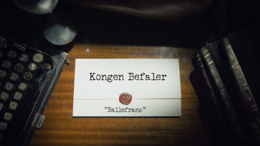 Image of the title card at the start of this episode, showing a task brief with the show title, 'Kongen Befaler', and the episode title, 'Ballefrans', on a wooden desk. At the edges of the image, part of a typewriter keyboard and a stack of leather-bound books can be seen.