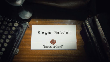 Image of the title card at the start of this episode, showing a task brief with the show title, 'Kongen Befaler', and the episode title, 'Pappa er her!' ['Daddy's here!'], on a wooden desk. At the edges of the image, part of a typewriter keyboard and a stack of leather-bound books can be seen.