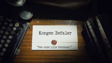 Image of the title card at the start of this episode, showing a task brief with the show title, 'Kongen Befaler', and the episode title, 'Den onde lille fetteren' ['The evil little cousin'], on a wooden desk. At the edges of the image, part of a typewriter keyboard and a stack of leather-bound books can be seen.