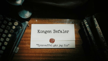 Image of the title card at the start of this episode, showing a task brief with the show title, 'Kongen Befaler', and the episode title, 'Tynnvaffel går jeg for' ['I'm going for a thin waffle'], on a wooden desk. At the edges of the image, part of a typewriter keyboard and a stack of leather-bound books can be seen.