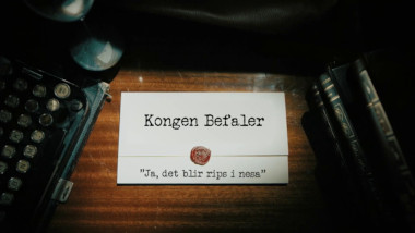 Image of the title card at the start of this episode, showing a task brief with the show title, 'Kongen Befaler', and the episode title, 'Ja, det blir rips i nesa' ['Yeah, it’ll be redcurrants in the nose'], on a wooden desk. At the edges of the image, part of a typewriter keyboard and a stack of leather-bound books can be seen.