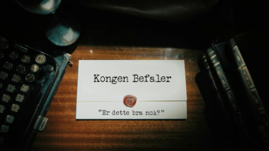 Image of the title card at the start of this episode, showing a task brief with the show title, 'Kongen Befaler', and the episode title, 'Er dette bra nok?' ['Is this good enough?'], on a wooden desk. At the edges of the image, part of a typewriter keyboard and a stack of leather-bound books can be seen.