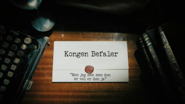 Image of the title card at the start of this episode, showing a task brief with the show title, 'Kongen Befaler', and the episode title, 'Men jeg men men den er vel er den ja' ['But I but but yeah well it's that one then'], on a wooden desk. At the edges of the image, part of a typewriter keyboard and a stack of leather-bound books can be seen.