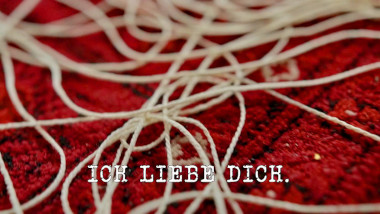 Image of a tangle of string on a red carpet (a reference to the 'Put a roll of string on the cushion' task), with the episode title, 'Ich liebe dich', superimposed on it.