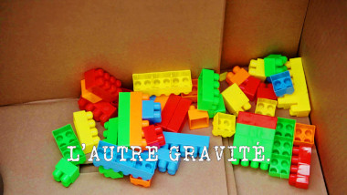 Image of some Duplo bricks in a cardboard box (a reference to the 'Get the most eggs into a frying pan' task, from the previous episode), with the episode title, 'L'autre gravité' ['The other gravity'], superimposed on it.