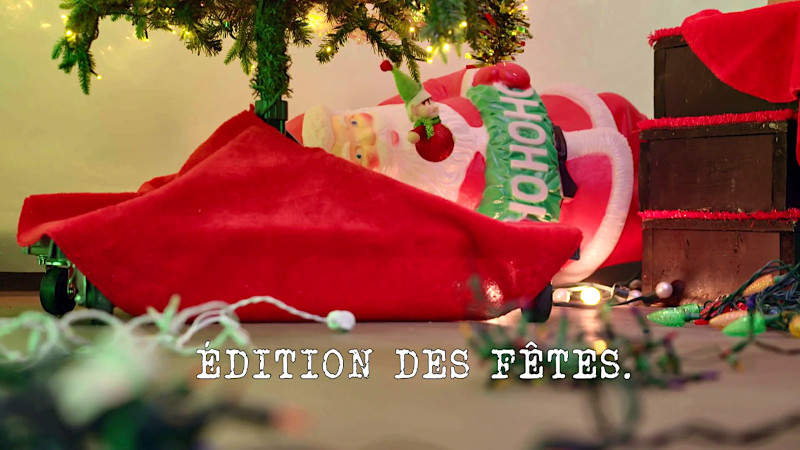 Image of the base of a Christmas tree, covered with red fabric, against which a plastic Santa Claus figurine is laid, while there are Christmas lights scattered across the floor in the foreground (a reference to the 'Untangle the Christmas lights' task) with the episode title 'Edition des Fêtes' [Holiday Special] superimposed on it.