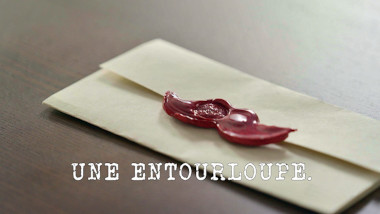 Image of a task brief laid on a table surface, with a wax seal in the shape of a moustache (a reference to the 'Make a moustache out of something unexpected' task), with the episode title 'Une entourloupe' [A scam] superimposed on it.