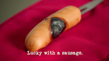 Image of a burned sausage on the end of a fork (a reference to the 'Save the snag from the barbie' task), with the episode title, 'Lucky with a sausage', superimposed on it.
