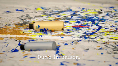 Image of a canvas drop-cloth spattered with paint, and with two bottles of poster paint laid upon it (a reference to the 'Paint an accurate map of Australia' task), with the episode title, 'Dumb in unison', superimposed on it.