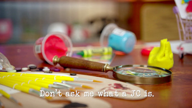 Image of a desk covered in craft supplies, and a magnifying glass (a reference to the 'Make a miniature version of yourself' task), with the episode title, 'Don't ask me what a JC is', superimposed on it.