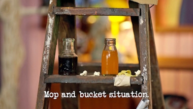 Image of some bottles with different beverages in them, balanced on the steps of a shabby wooden step-ladder (a reference to Aaron Chen's attempt at the 'Turn yourself into a piece of furniture' task), with the episode title, 'Mop and bucket situation', superimposed on it.