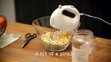 Image of an electric whisk balanced in a glass bowl of hard sweets on a kitchen counter (a reference to Aaron Chen's attempt at the ‘Make the most obnoxiously loud racket with a racquet’ task), with the episode title, ‘A bit of a pickle’, superimposed on it.