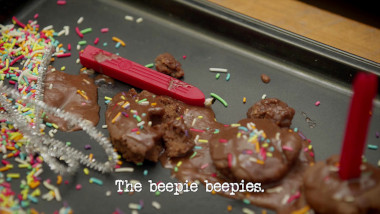 Image of some small chocolate cakes on a baking tray, with hundreds and thousands unevenly sprinkled over them, and small red sealing wax candles instead of normal birthday candles (a reference to Emma Holland's attempt at the 'Celebrate your selected national day' task), with the episode title 'The beepie beepies' superimposed on it.