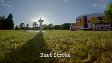 Image of a backlit plinth in front of a tarpaulin, near the Taskmaster caravan (a reference to the 'Squirt sunscreen the furthest' task), with the episode title, 'Heat stroke', superimposed on it.