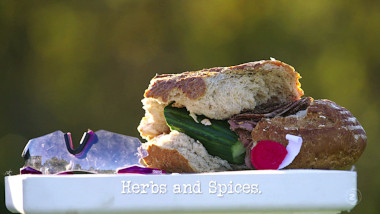 Image of a badly made sandwich and some safety goggles on the top of a plinth (a reference to the 'Drive 11 laps and obey all the rules' task), with the episode title, 'Herbs & Spices', superimposed on it.