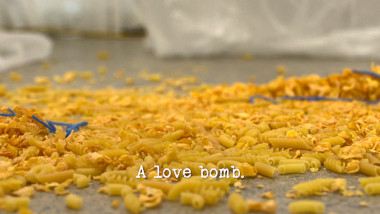 Image of a mixture of dry goods, including pasta and breakfast cereal, scattered on the floor of the lab of the Taskmaster house (a reference to Dai Henwood's attempt at the 'Do the most unpredictable thing' task), with the episode title, 'A love bomb', superimposed on it.