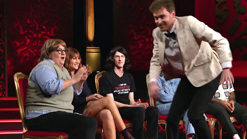 Image of Taskmaster AU’s assistant Tom Cashman making his cameo appearance as Paul William’s stunt double, running back across the stage in front of the contestants, who are all wearing wigs.