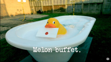 Image of a rubber duck holding a task in its bill, floating in the bathtub full of soapy water in the TaskMaster garden (taken during the 'Empty the bathtub' task), with the episode title, ‘Melon buffet’, superimposed on it.
