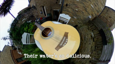 A 360 camera image of an acoustic guitar on a garden table (a reference to the 'Write and perform a song about a stranger' task), with the episode title, 'Their water’s so delicious', superimposed on it.