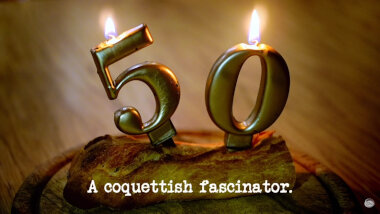 Image of lit novelty number-shaped 50th birthday candles, stuck into a piece of a baguette (a reference to it being the 50th episode of the show), with the episode title, 'A coquettish fascinator', superimposed on it.